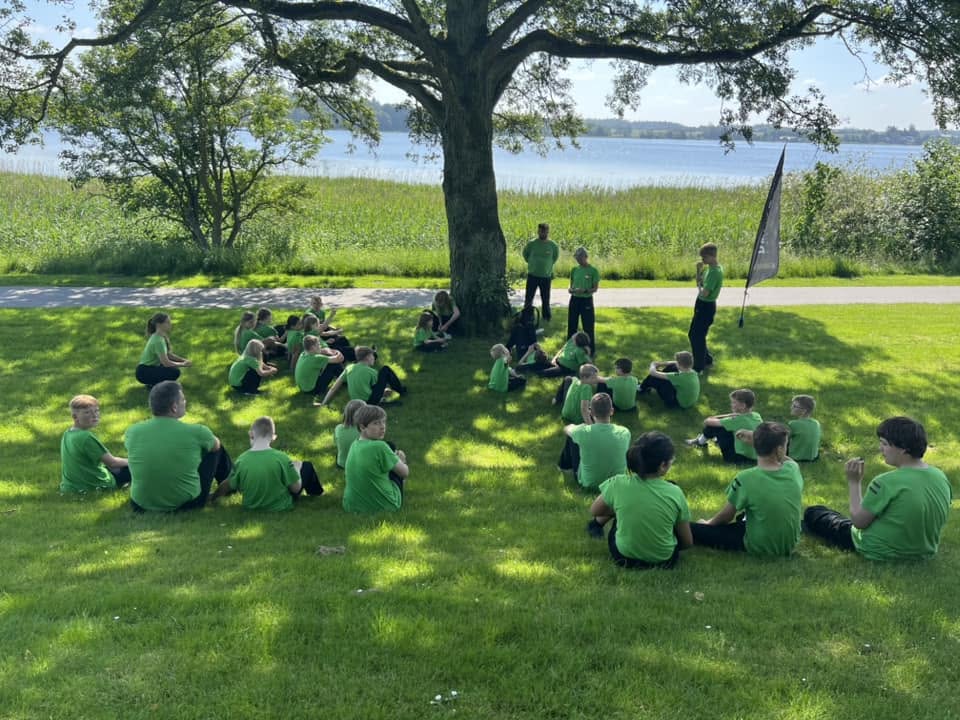 Børn fra Dai Ki Haku sidder i græsset under et træ ved søen